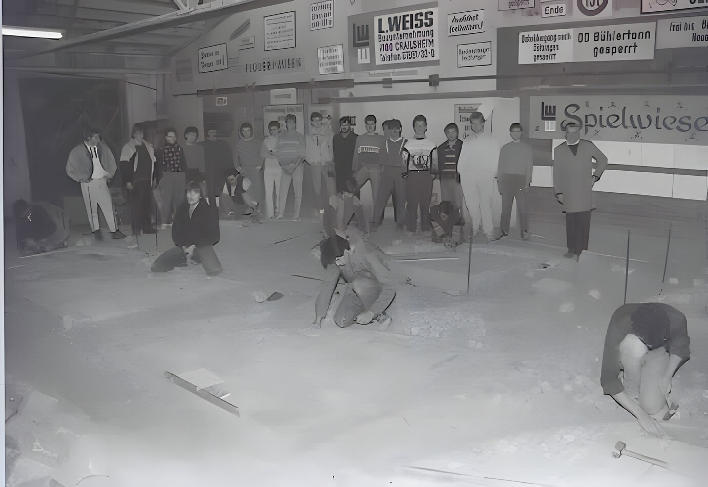 Schwarz-Weiß-Bild von sechs Auszubildenden, die in einer Halle Pflasterarbeiten ausführen, während eine Gruppe weiterer Personen dabei zusieht