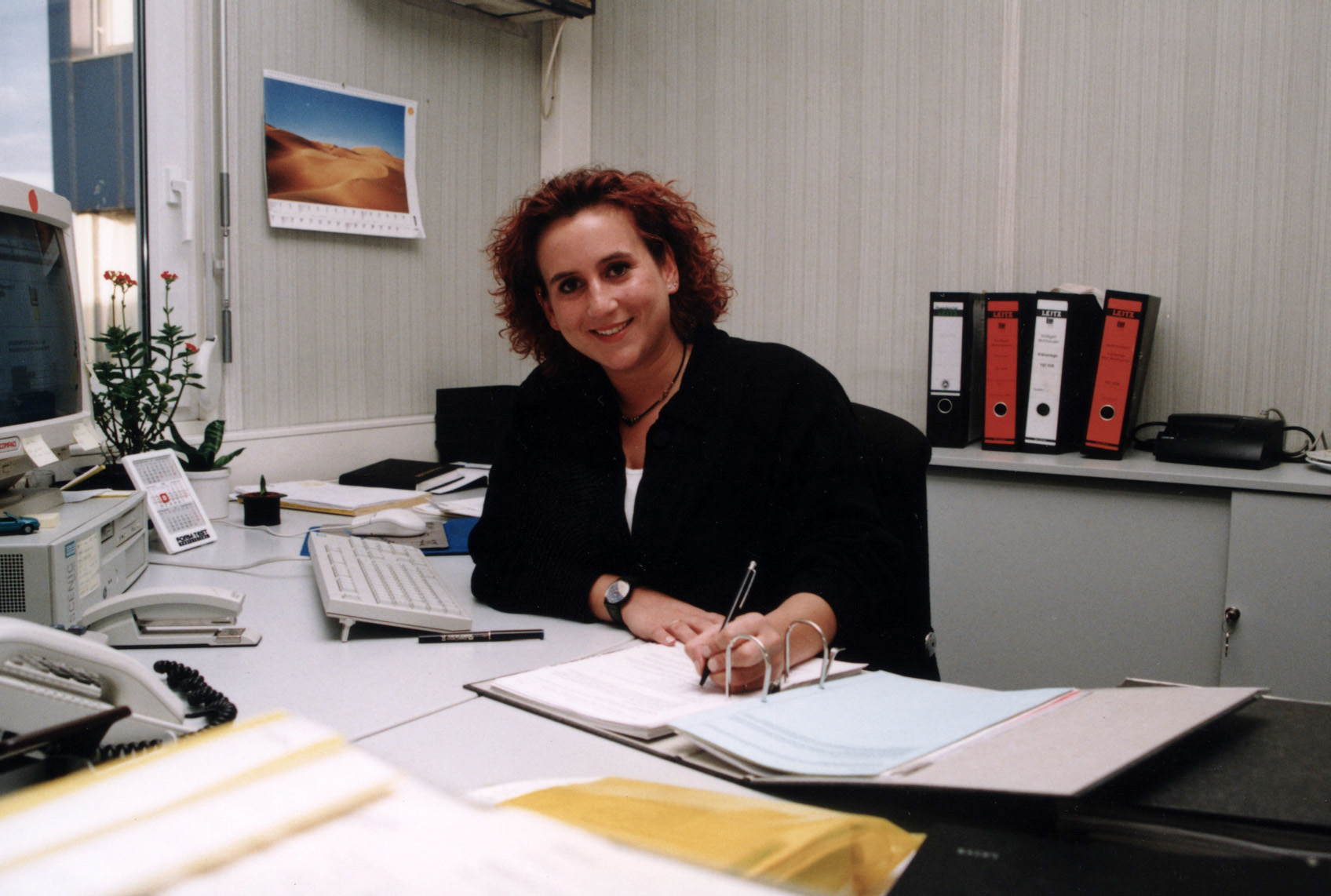 Farbfotografie einer jungen Frau mit roten Locken an einem Schreibtisch in einem Büro