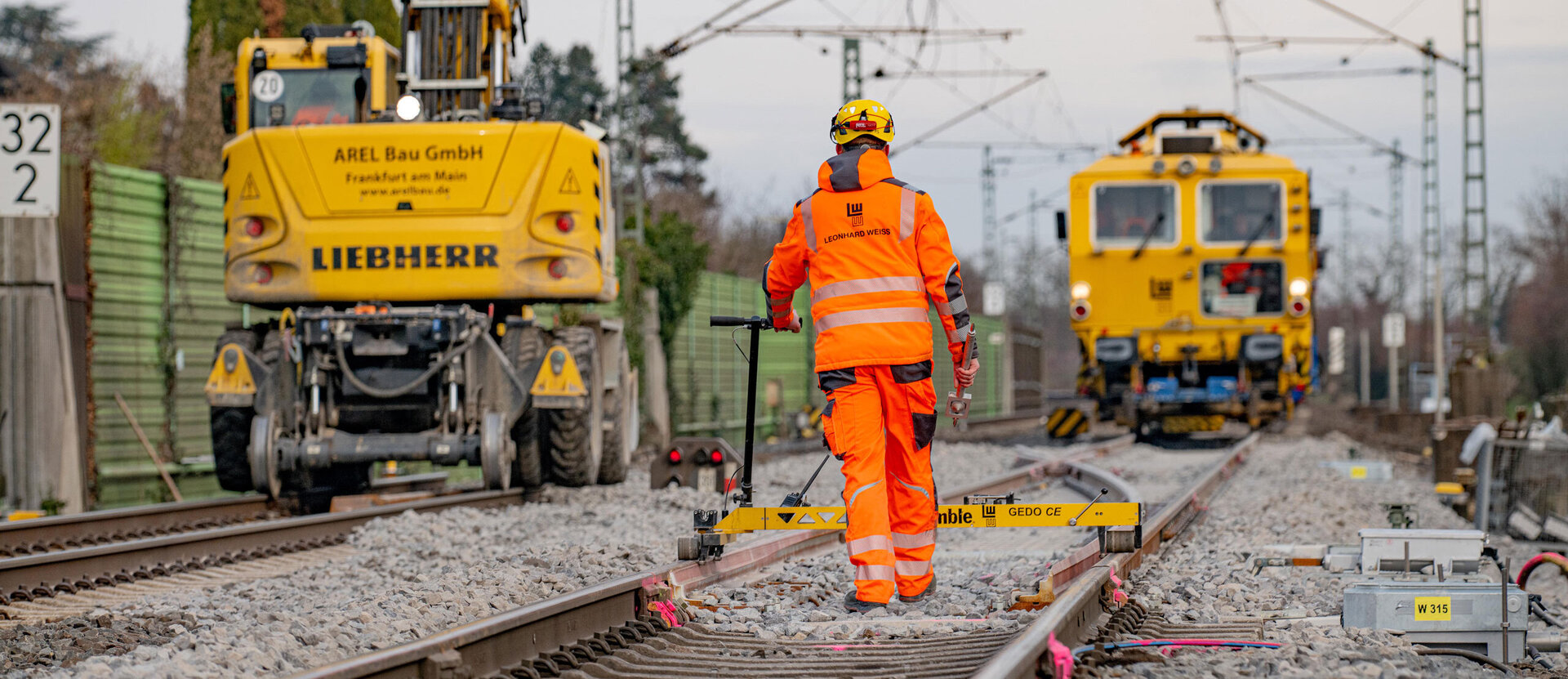 Bauarbeiten an der Riedbahn: Ein Mitarbeiter in orangener Schutzkleidung führt Vermessungen auf den Gleisen durch, im Hintergrund stehen gelbe Maschinen