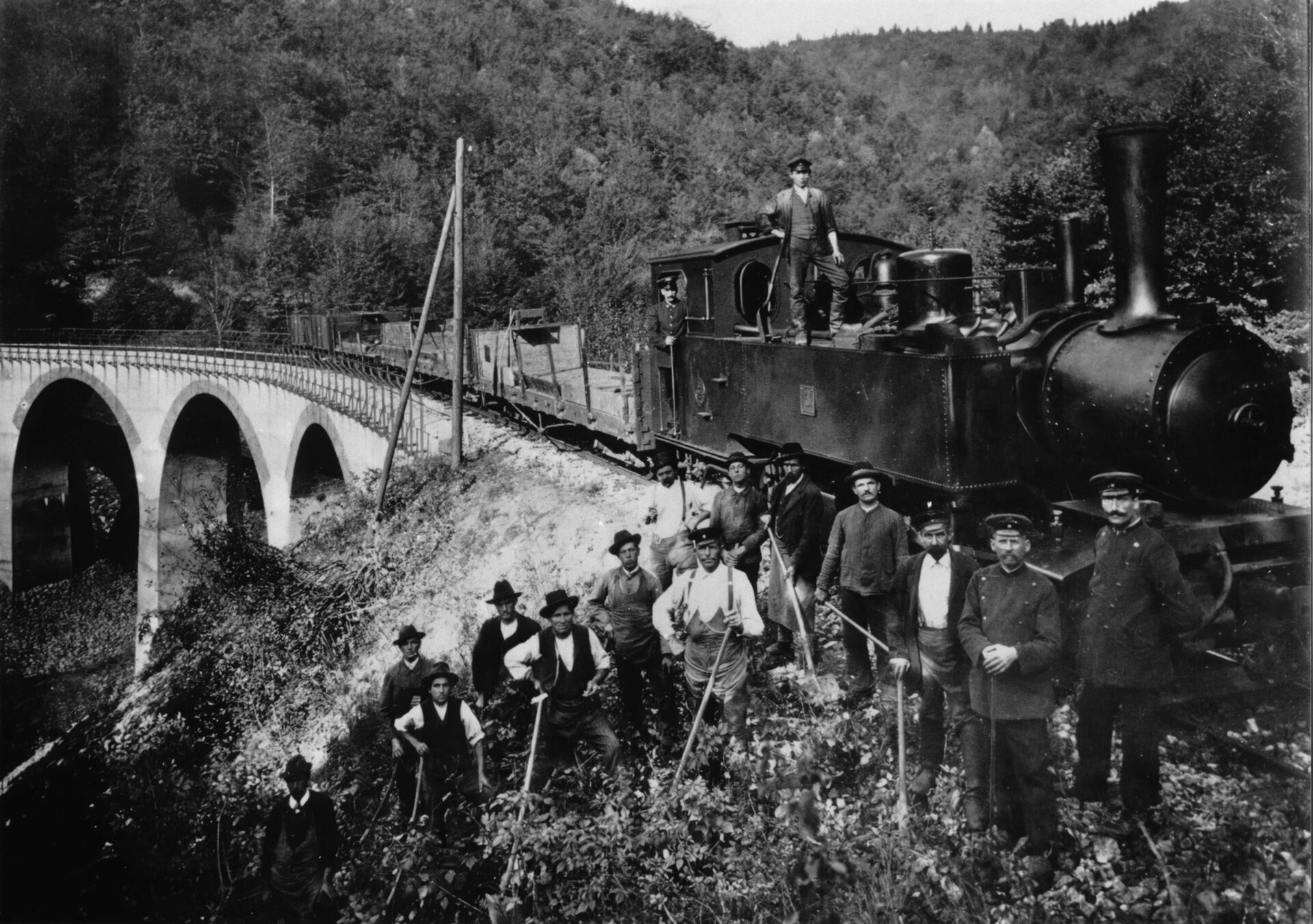 Schwarz-Weiß-Foto einer Lokomotive der Härtsfeldbahn mit Arbeitern und Bahnpersonal, aufgenommen vor einem Viadukt in einer waldreichen Umgebung