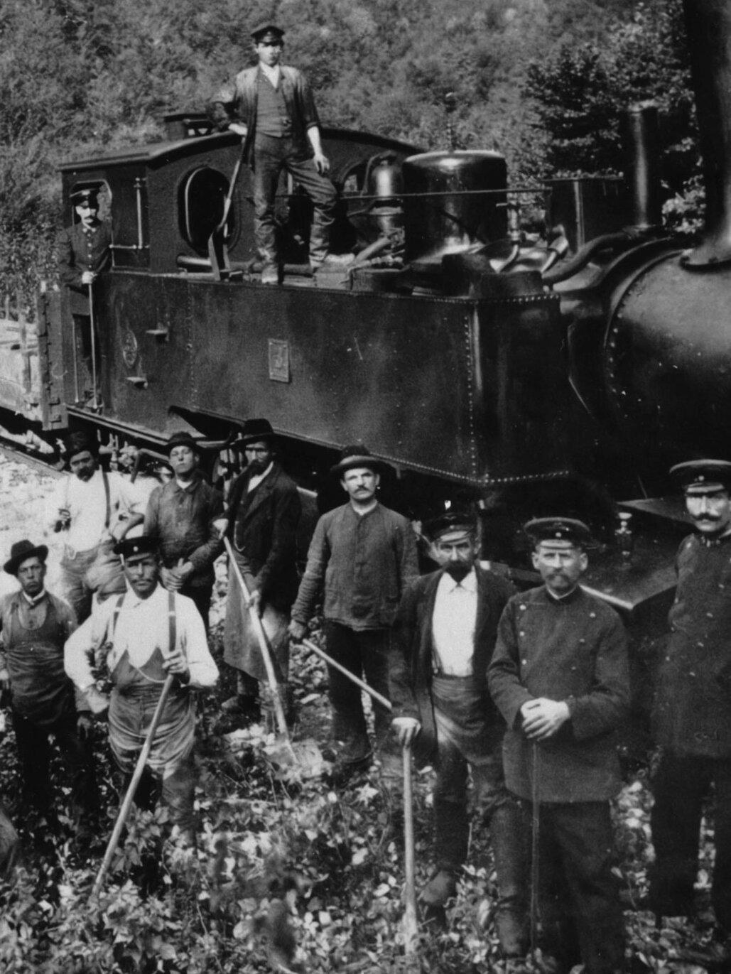 Schwarz-Weiß-Foto einer Lokomotive der Härtsfeldbahn mit Arbeitern und Bahnpersonal, aufgenommen vor einem Viadukt in einer waldreichen Umgebung