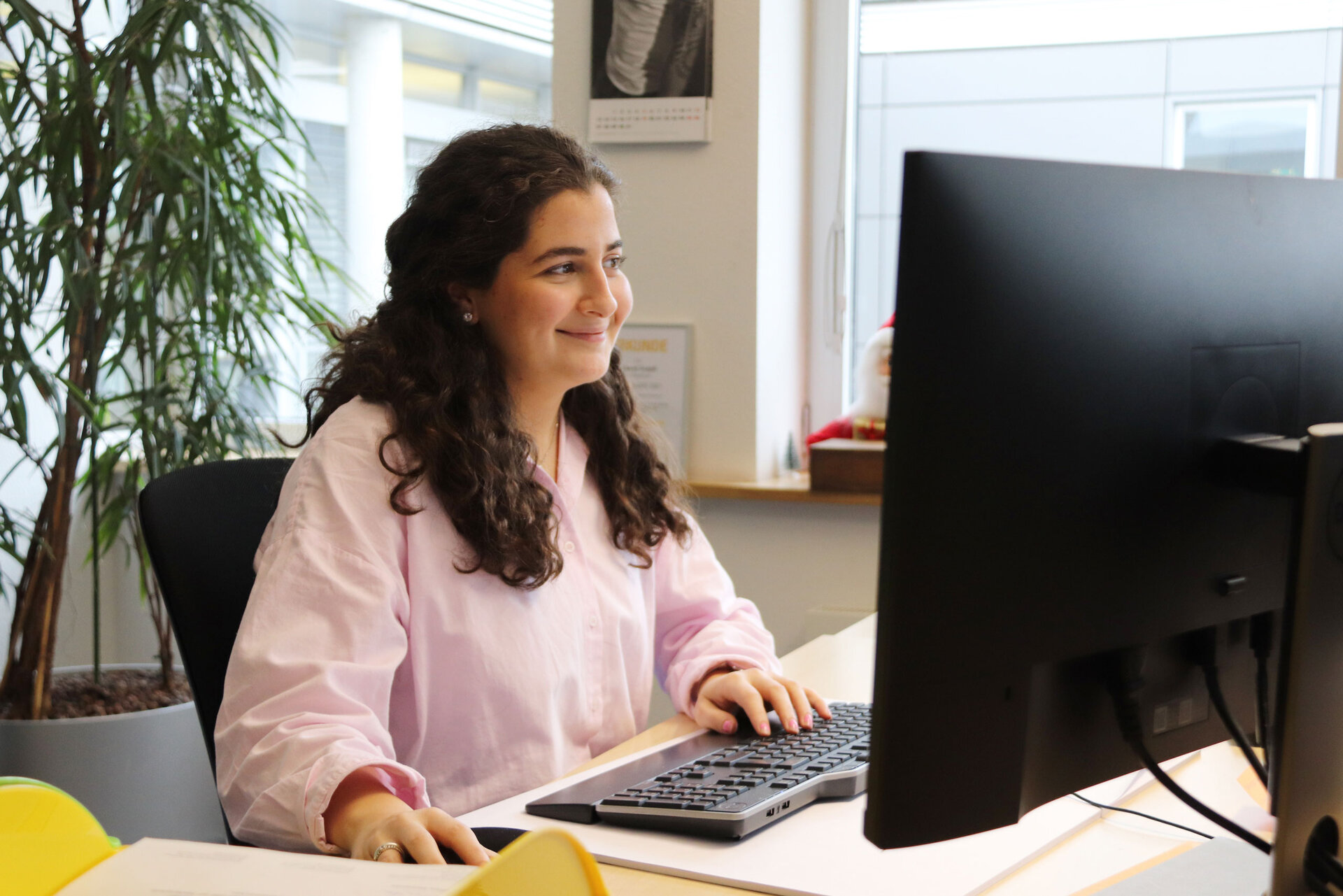 Junge Frau mit braunen Locken und rosa Bluse am Schreibtisch vor einem Computer, mit Pflanze und Fenster im Hintergrund