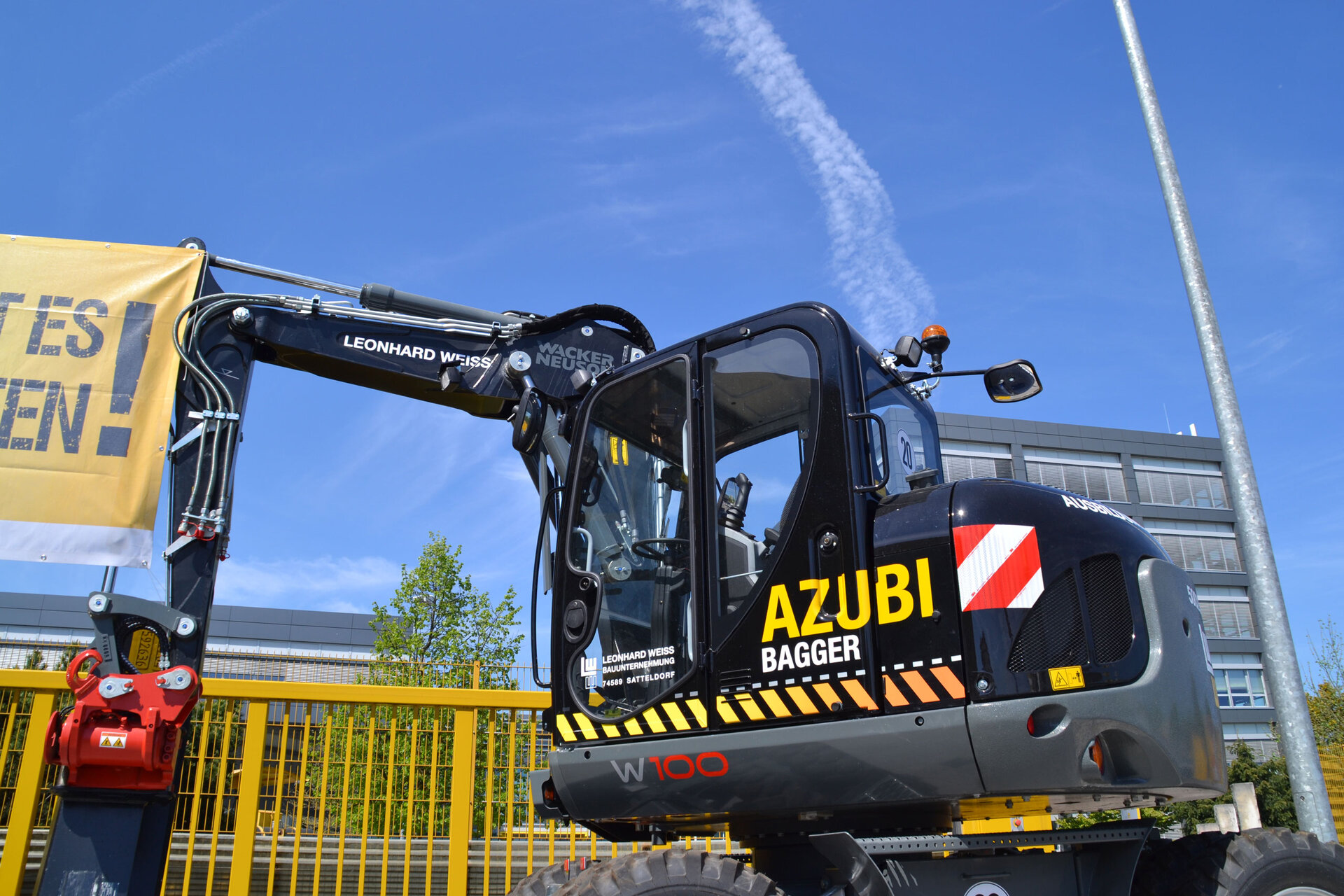 Schwarzer Bager mit der Aufschrift ‚AZUBI BAGGER‘ vor einem gelben Zaun und blauem Himmel im Hintergrund