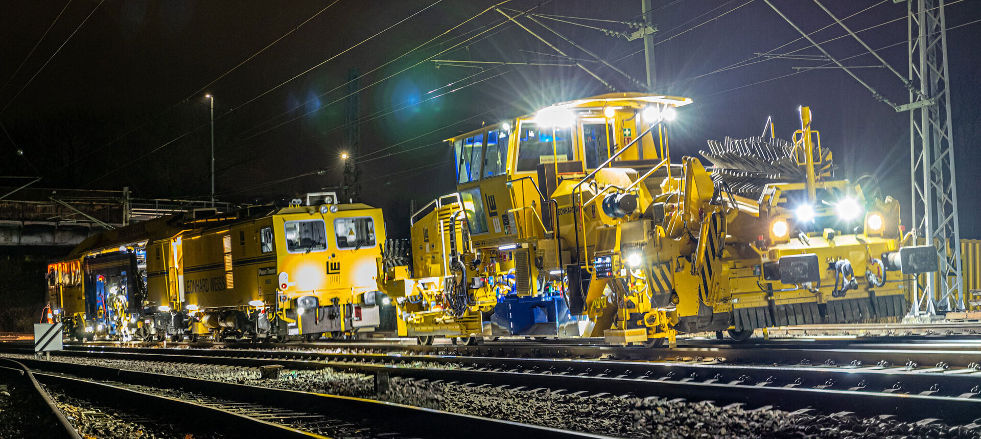 Gleisbauarbeiten bei Nacht mit beleuchteten gelben Maschinen auf einer elektrifizierten Bahnstrecke