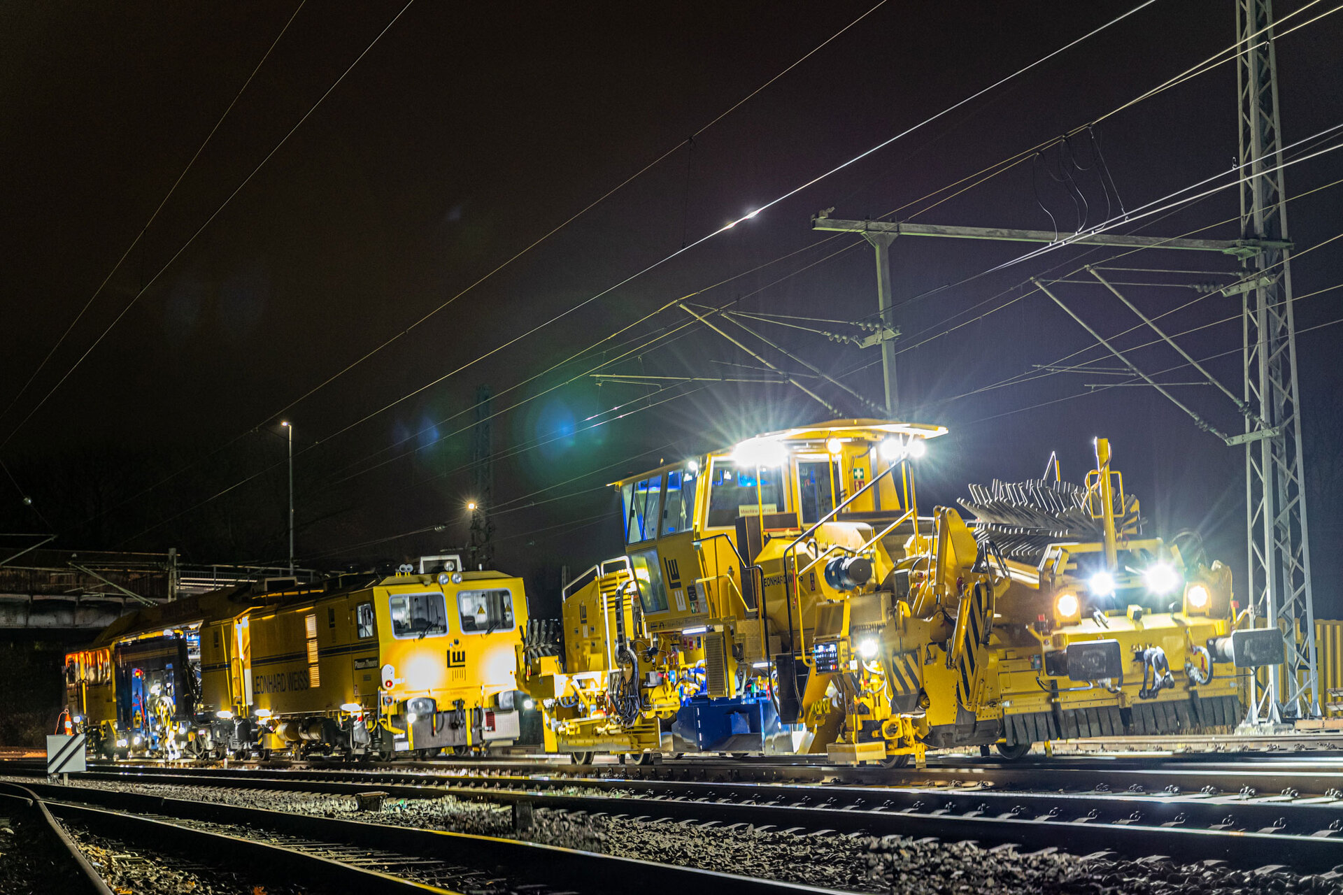 Gleisbauarbeiten bei Nacht mit beleuchteten gelben Maschinen auf einer elektrifizierten Bahnstrecke