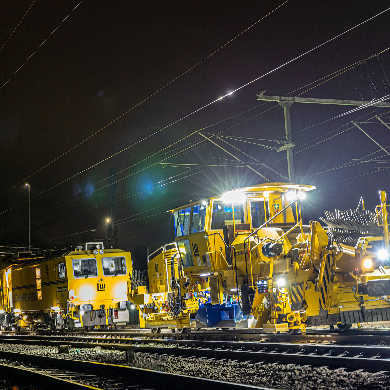 Track construction work at night with illuminated yellow machines on an electrified railway line