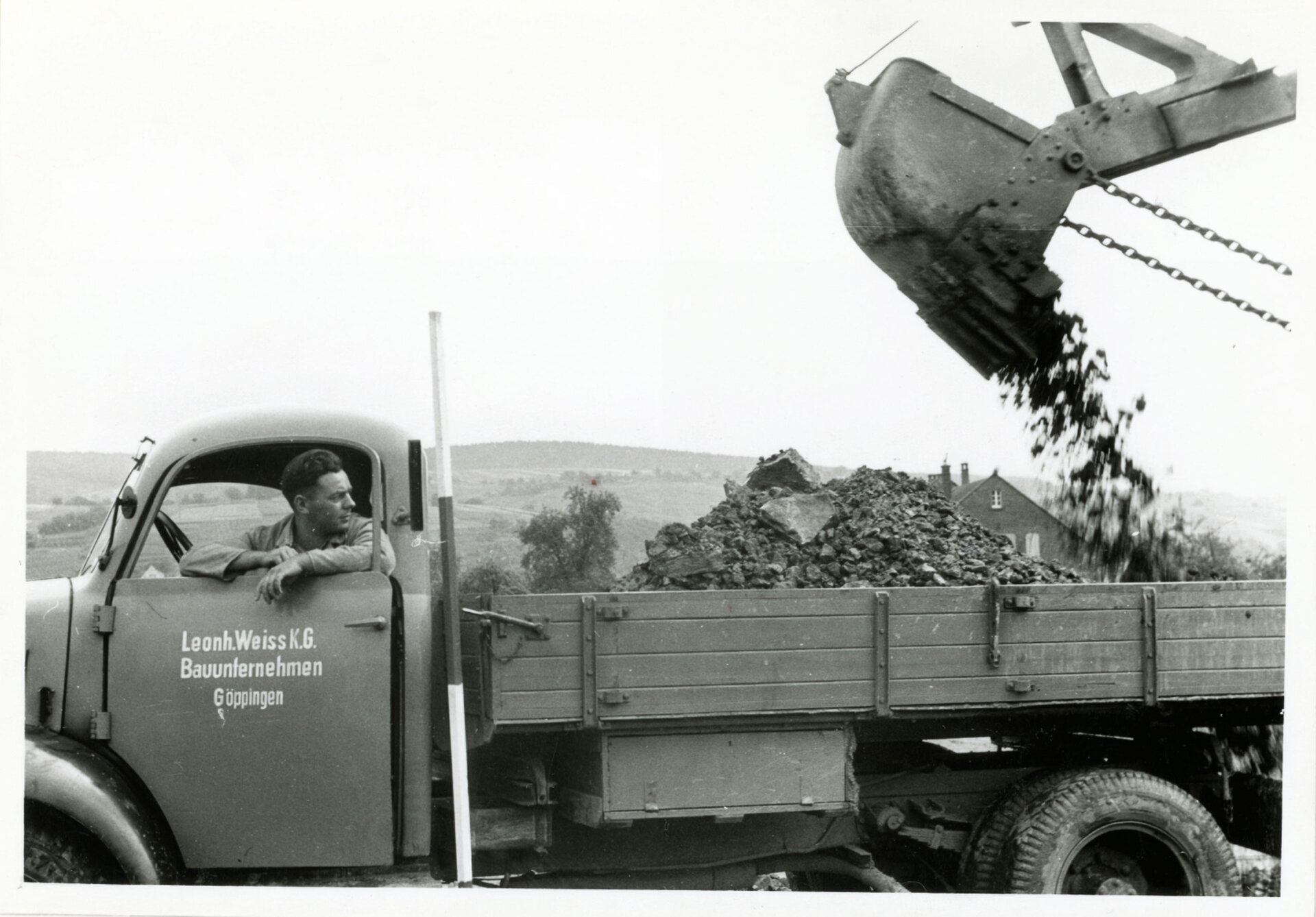 Schwarz-Weiß-Foto eines Lkw mit der Aufschrift 'Leonh. Weiss K.G. Bauunternehmen Göppingen', dessen Ladefläche mit einem Bagger mit Schutt beladen wird