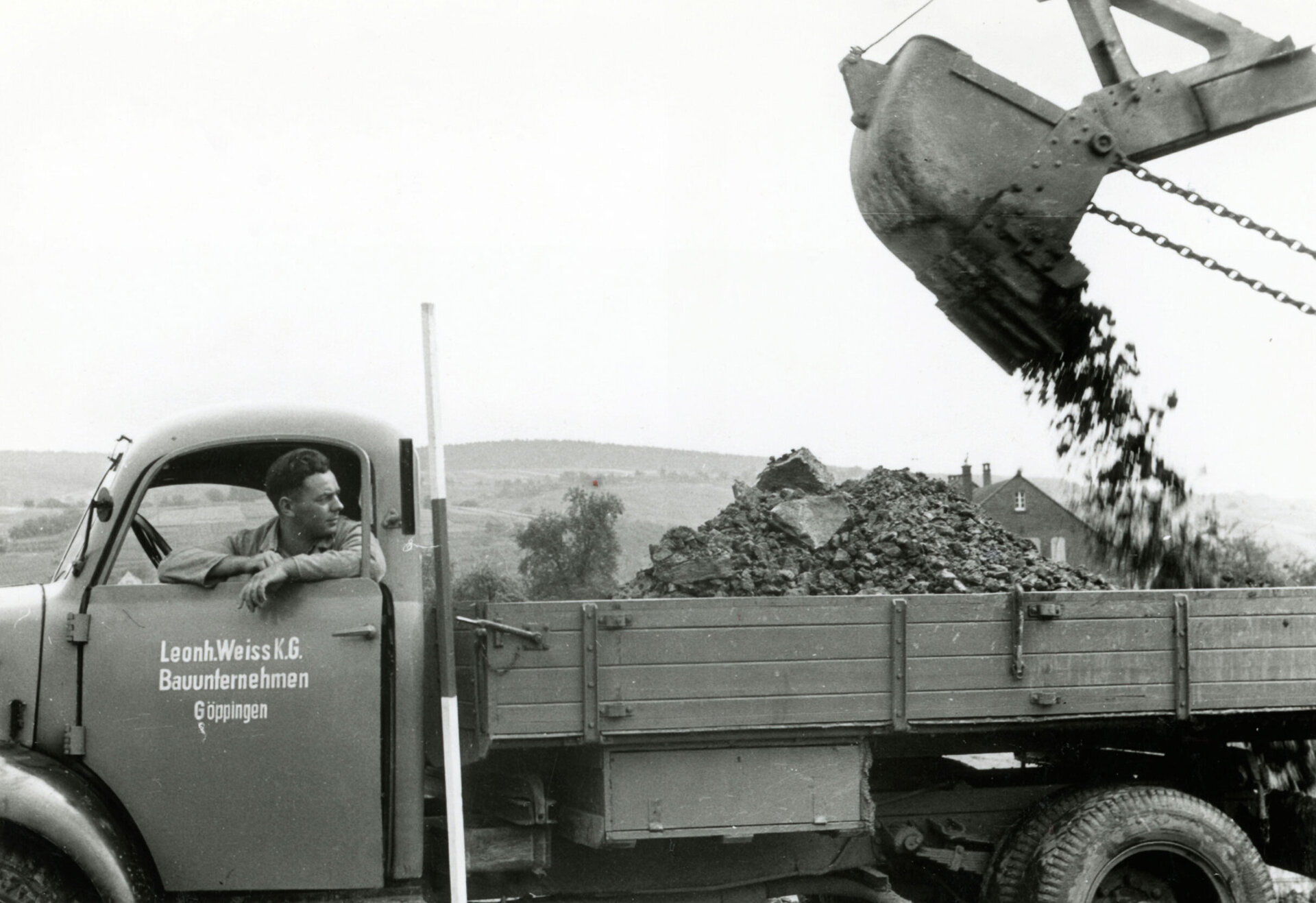 Schwarz-Weiß-Foto eines Lkw mit der Aufschrift 'Leonh. Weiss K.G. Bauunternehmen Göppingen', dessen Ladefläche mit einem Bagger mit Schutt beladen wird