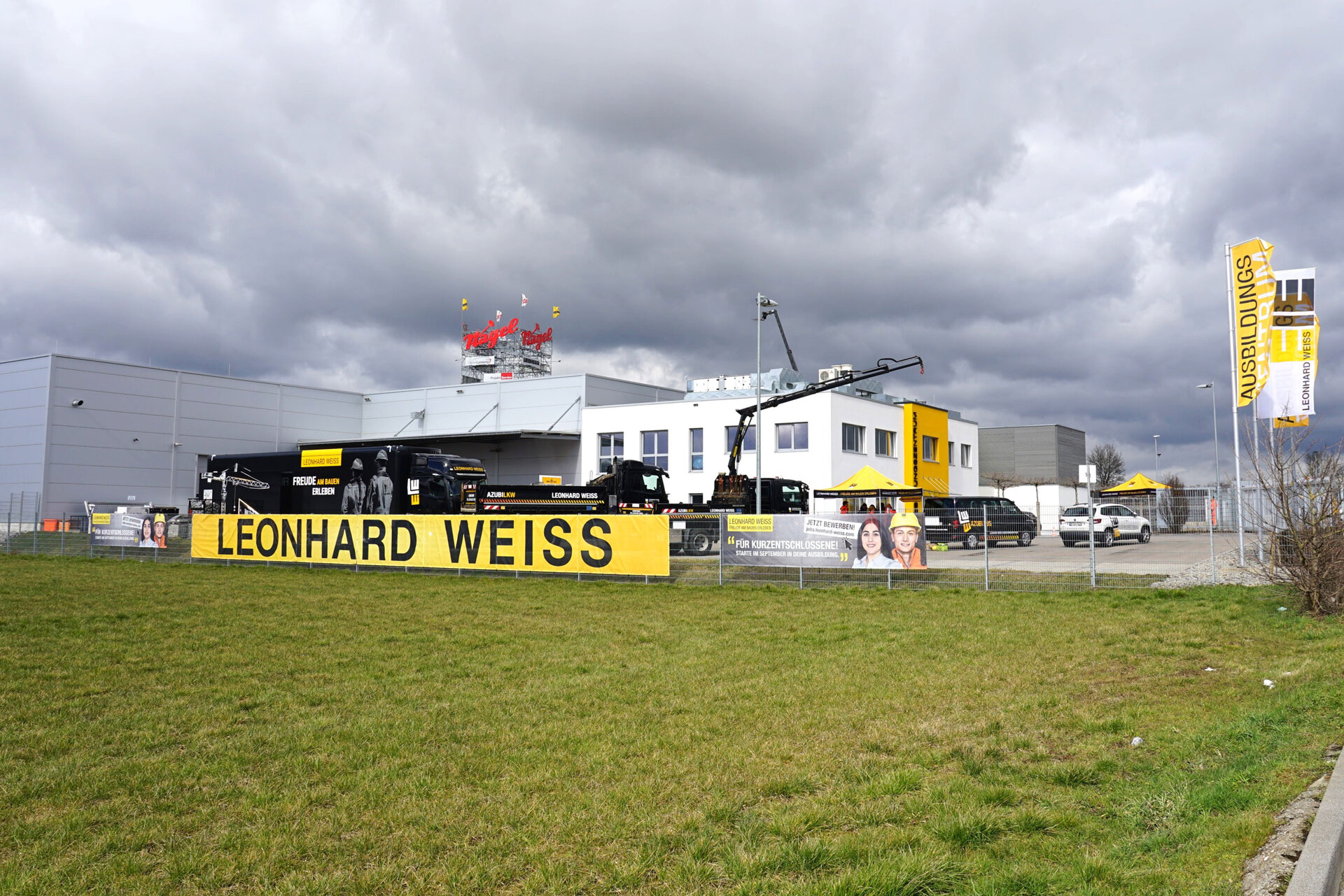 Ausbildungszentrum von LEONHARD WEISS mit gelber Beschilderung und Bannern, Wolkenhimmel im Hintergrund