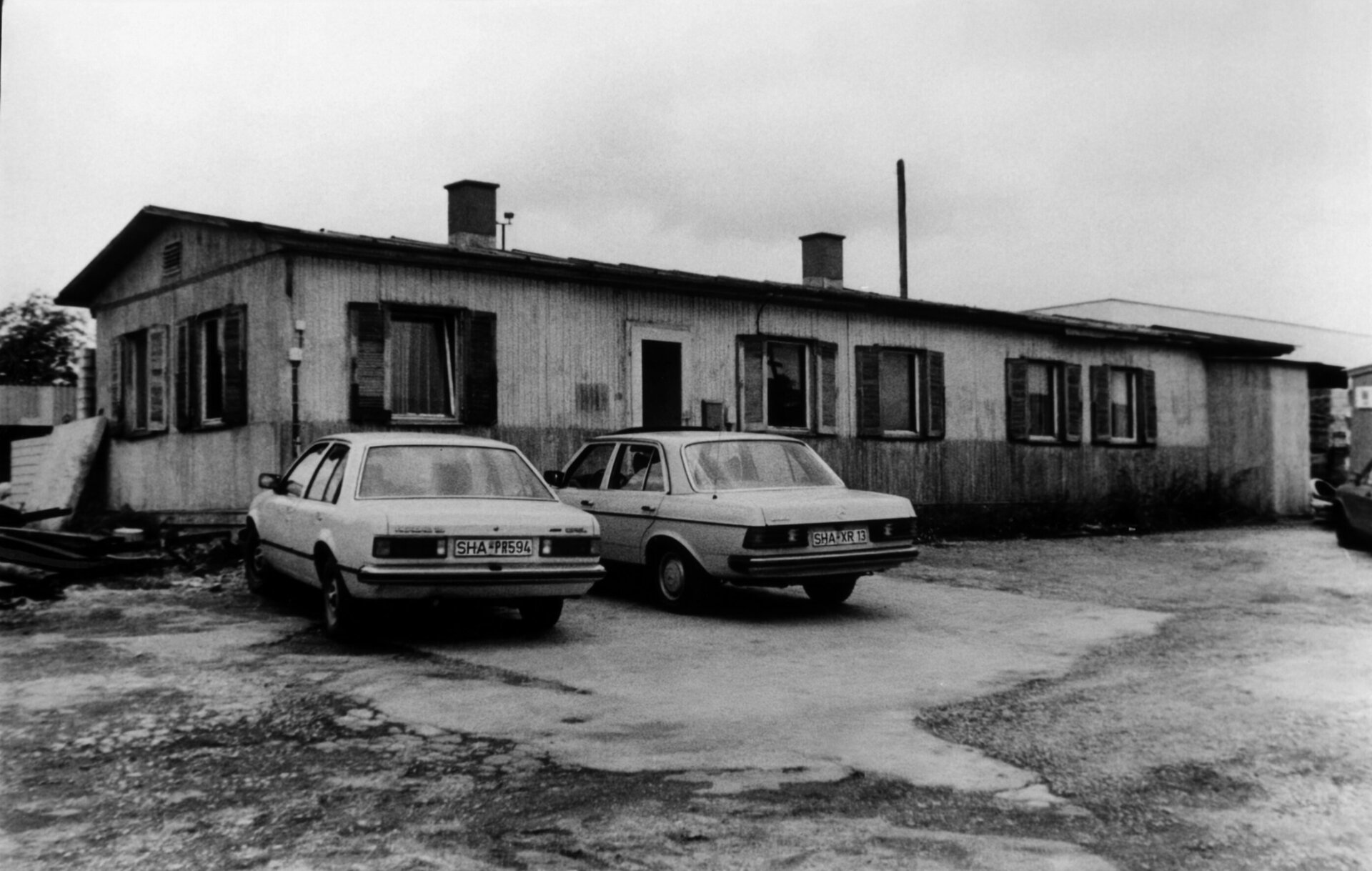 Schwarz-Weiß-Foto des LEONHARD WEISS Standorts in Crailsheim, mit zwei geparkten Autos vor einem einfachen Bürogebäude