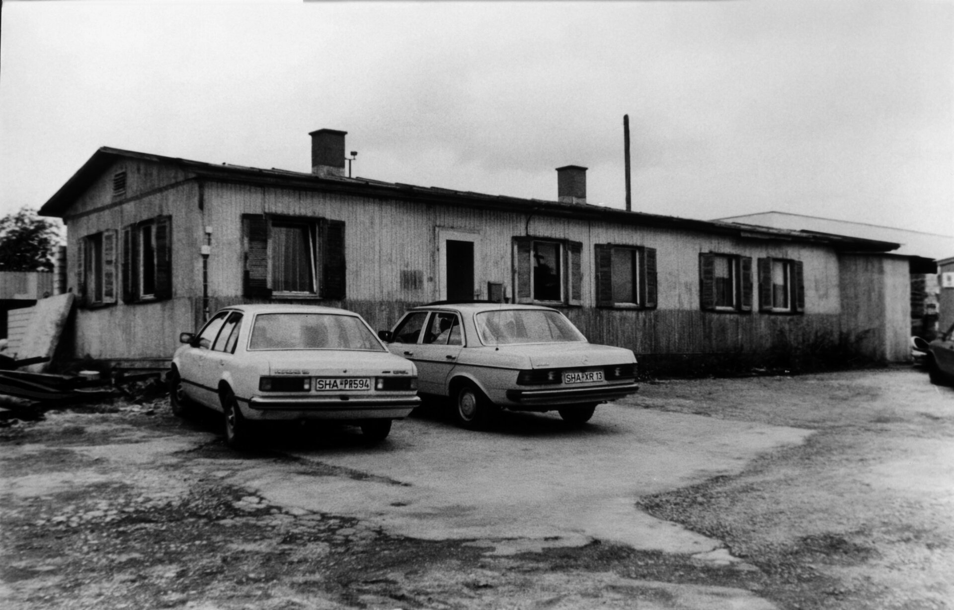 Schwarz-Weiß-Foto des LEONHARD WEISS Standorts in Crailsheim, mit zwei geparkten Autos vor einem einfachen Bürogebäude