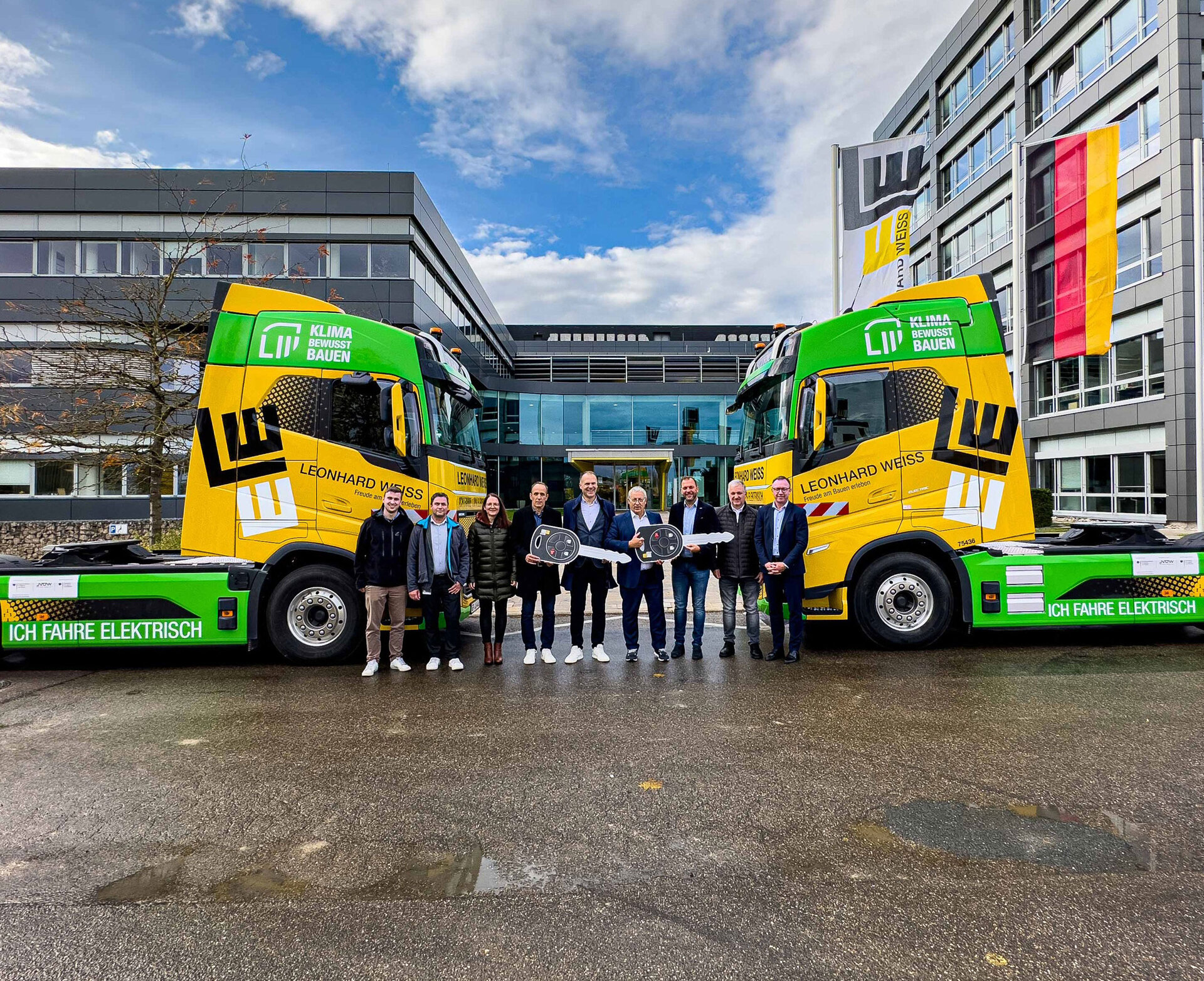 Zwei elektrisch betriebene LKW mit dem grünen Logo „KLIMA BEWUSST BAUEN“. Im Vordergrund stehen neun Personen, die symbolisch zwei große Schlüssel übergeben