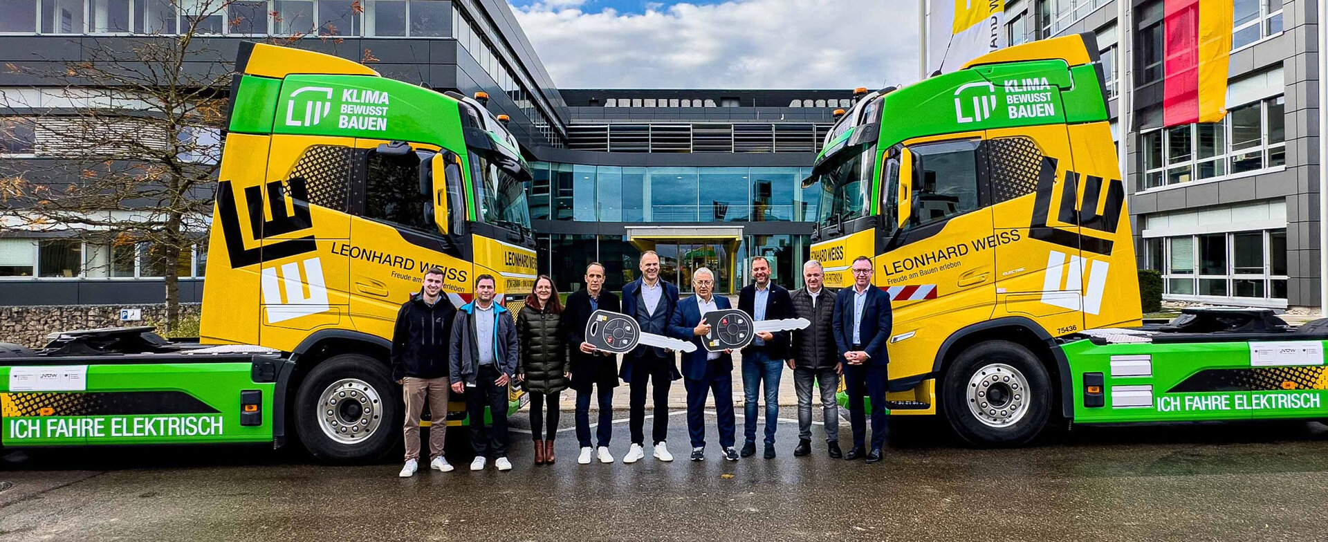 Zwei elektrisch betriebene LKW mit dem grünen Logo „KLIMA BEWUSST BAUEN“. Im Vordergrund stehen neun Personen, die symbolisch zwei große Schlüssel übergeben