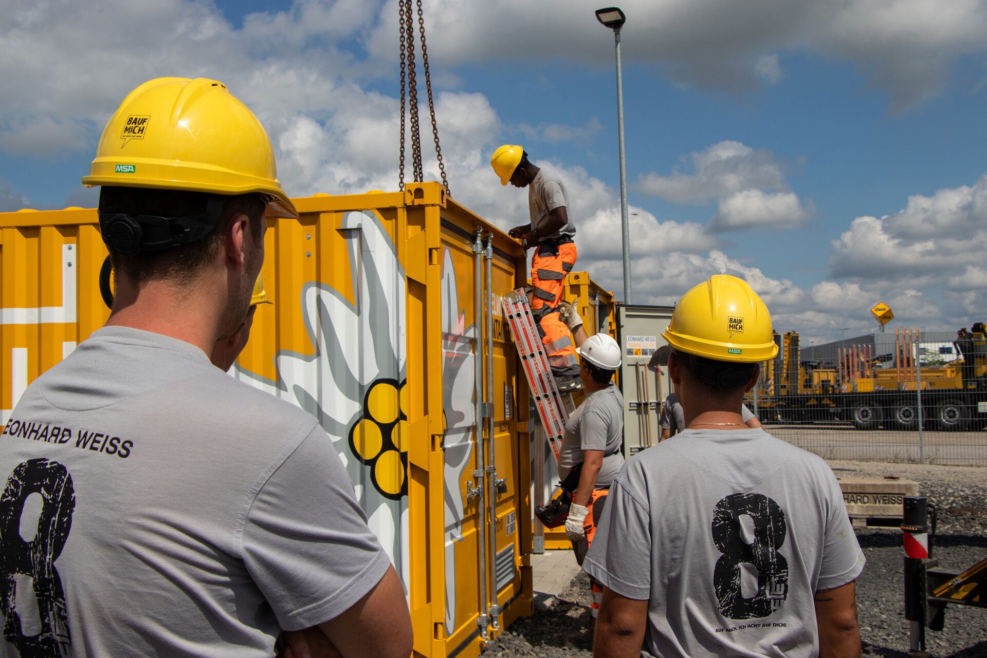 Mitarbeitende von LEONHARD WEISS mit gelben Helmen und grauen Shirts arbeiten an einem gelben Container auf einer Baustelle