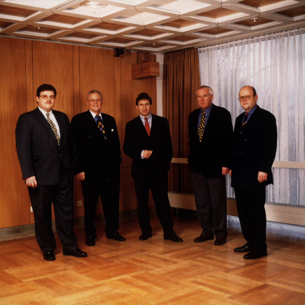 Gruppenfoto von fünf Männern in Anzügen, aufgenommen 1998 anlässlich des Zusammenschlusses der Standorte, in einem holzvertäfelten Raum