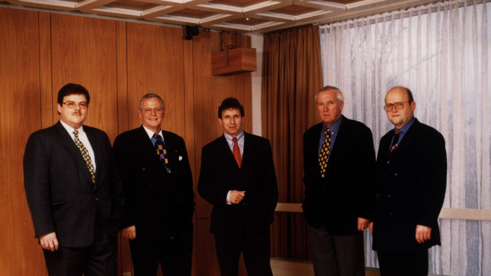 Gruppenfoto von fünf Männern in Anzügen, aufgenommen 1998 anlässlich des Zusammenschlusses der Standorte, in einem holzvertäfelten Raum