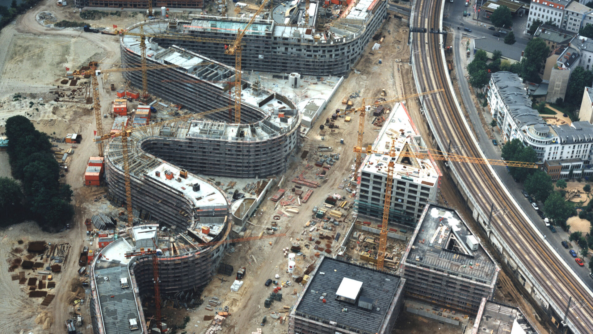 Luftaufnahme der Baustelle Moabiter Werder in Berlin. Mehrere Gebäude in verschiedenen Bauphasen, umgeben von Baukränen, einer Bahnstrecke und städtischer Infrastruktur