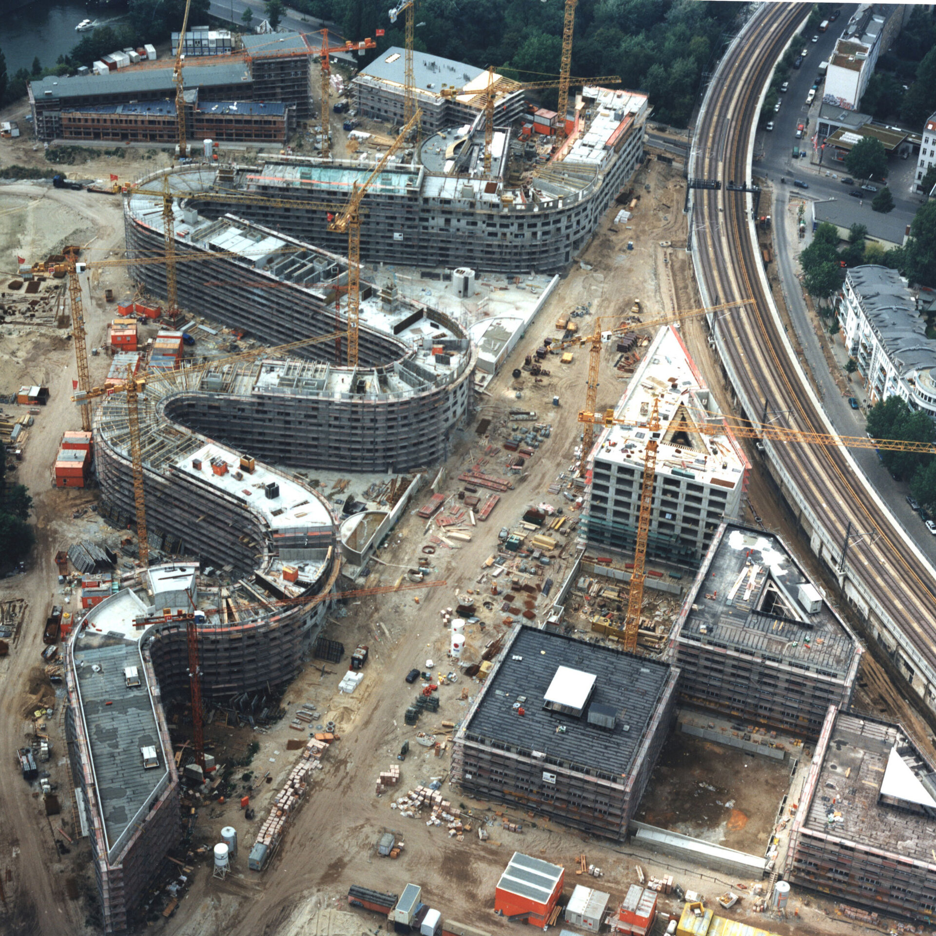 Luftaufnahme der Baustelle Moabiter Werder in Berlin. Mehrere Gebäude in verschiedenen Bauphasen, umgeben von Baukränen, einer Bahnstrecke und städtischer Infrastruktur