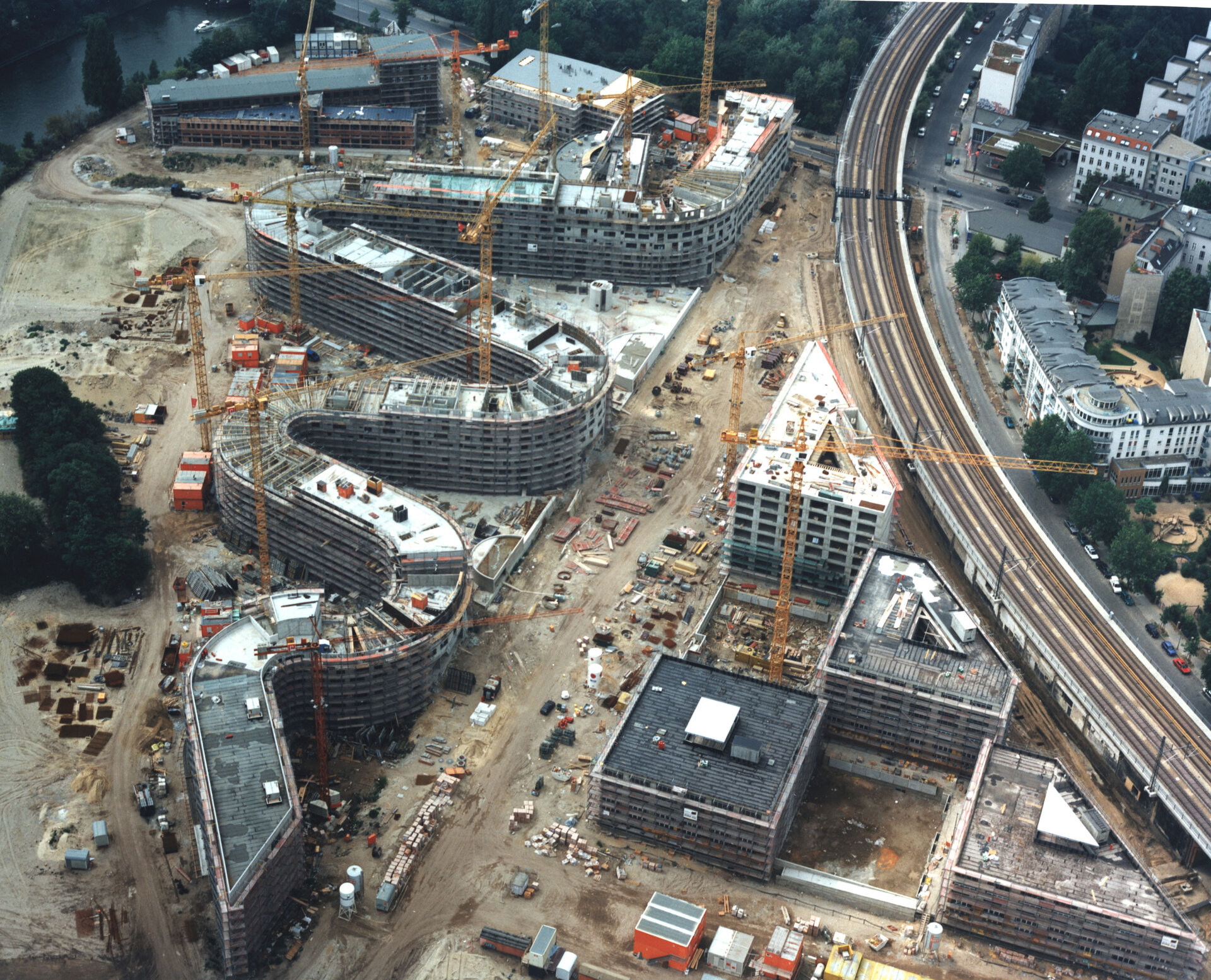 Luftaufnahme der Baustelle Moabiter Werder in Berlin. Mehrere Gebäude in verschiedenen Bauphasen, umgeben von Baukränen, einer Bahnstrecke und städtischer Infrastruktur
