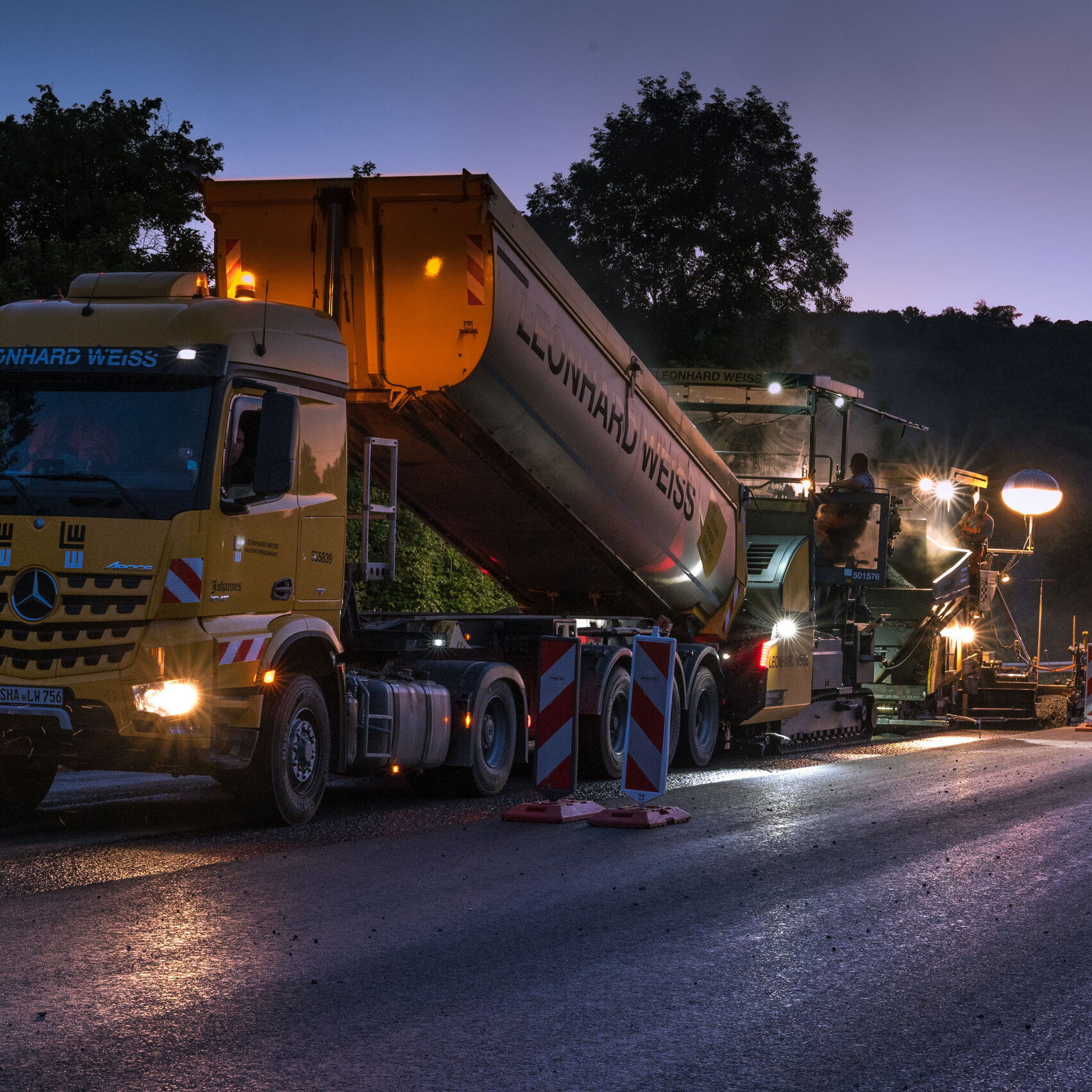 Night shot of a road construction project with yellow LEONHARD WEISS lorry and asphalting machine, illuminated by construction site lamps