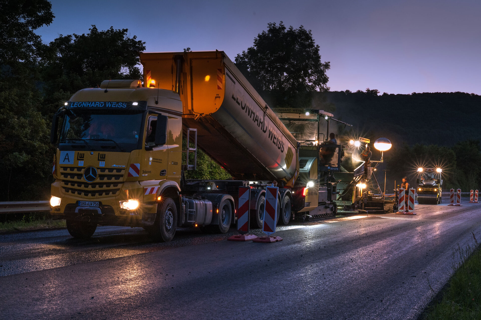 Nachtaufnahme eines Straßenbauprojekts mit gelbem LEONHARD WEISS LKW und Asphaltiermaschine, beleuchtet durch Baustellenlampen