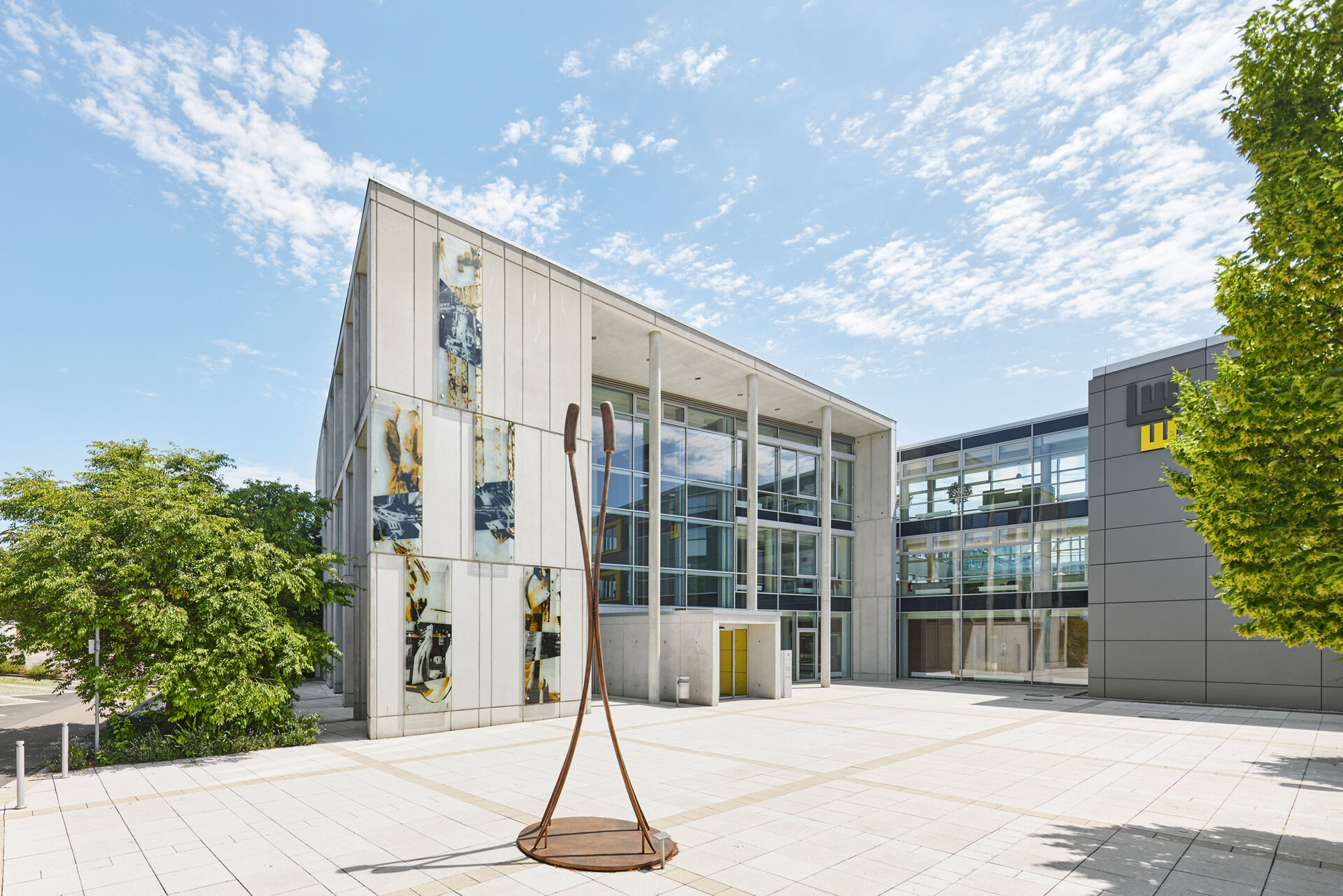 Moderner Gebäudekomplex des LEONHARD WEISS Standorts in Göppingen mit Glasfassaden, künstlerischen Betonwänden und einer Skulptur auf einem sonnigen Platz