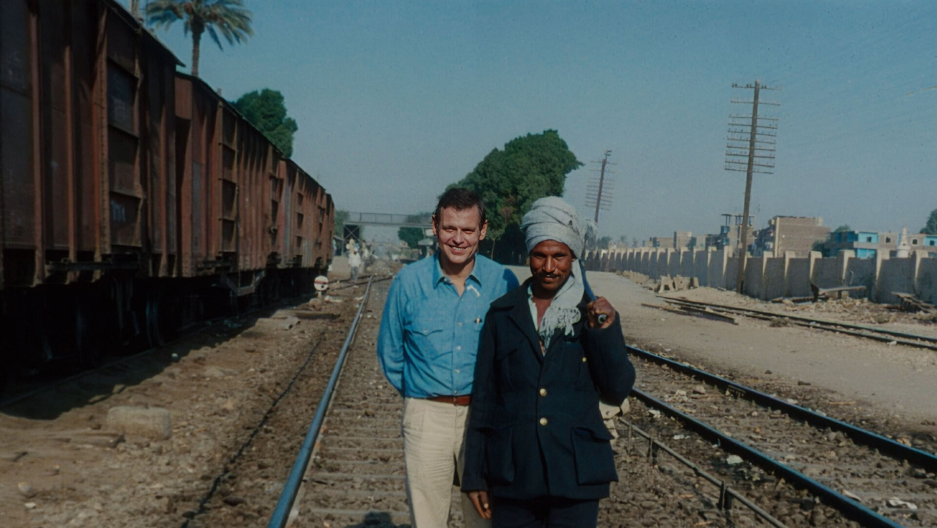 Zwei Männer stehen auf Bahngleisen in Ägypten, im Hintergrund Güterwaggons, Palmen und Gebäude