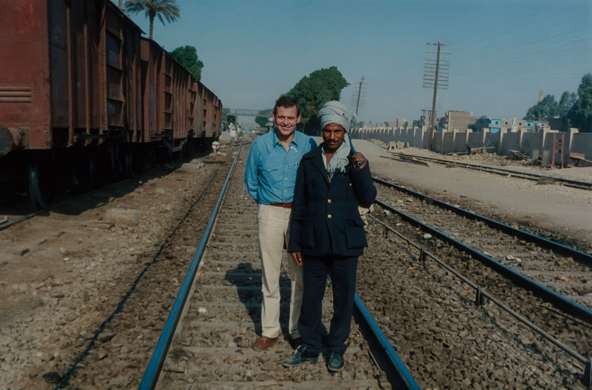 Zwei Männer stehen auf Bahngleisen in Ägypten, im Hintergrund Güterwaggons, Palmen und Gebäude