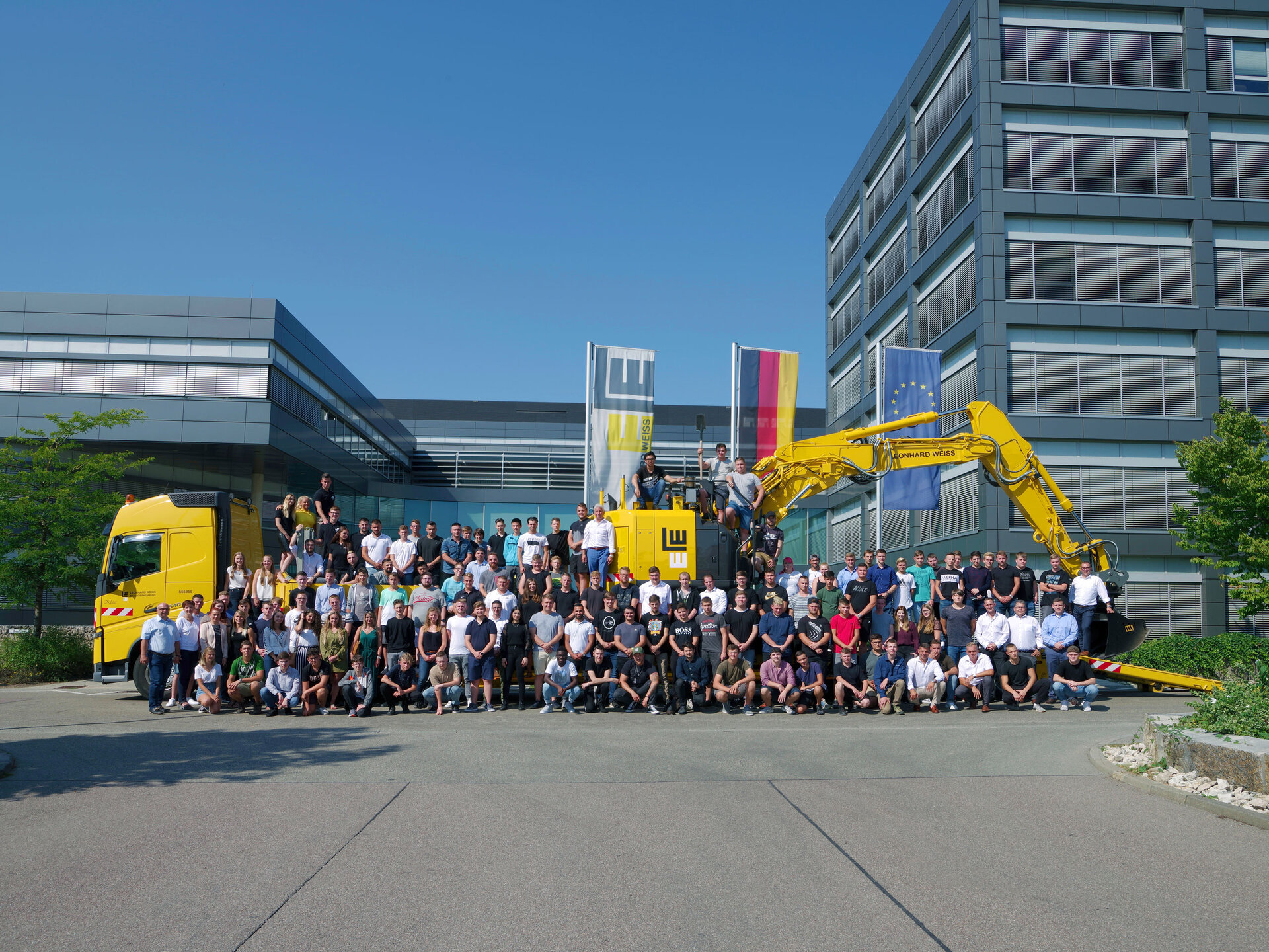 Gruppenfoto von Auszubildenden und Mitarbeitenden von LEONHARD WEISS vor einem gelben LKW, einem gelben Bagger und einem Bürogebäude