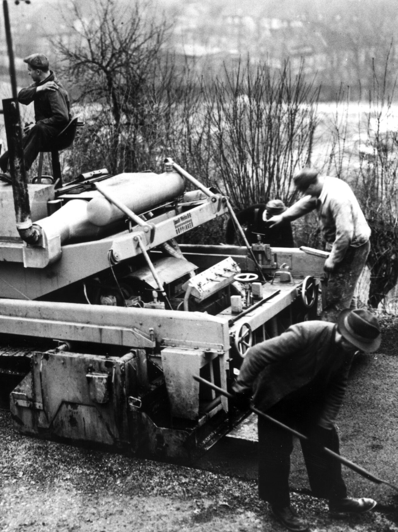 Schwarz-Weiß-Foto einer Straßenbaumaschine im Einsatz 1957, Arbeiter bearbeiten den Belag, im Hintergrund Gebäude und hügelige Landschaft