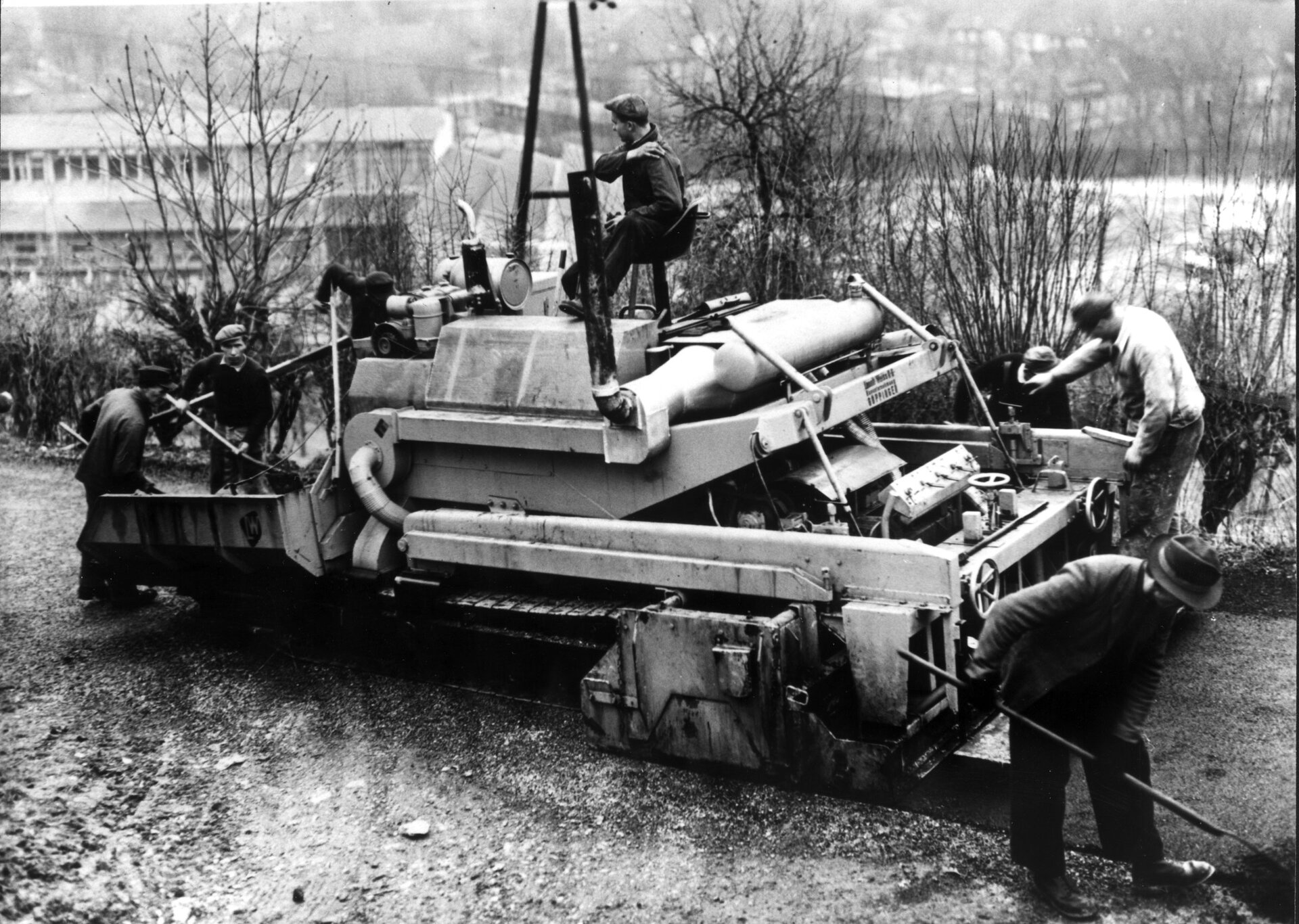 Schwarz-Weiß-Foto einer Straßenbaumaschine im Einsatz 1957, Arbeiter bearbeiten den Belag, im Hintergrund Gebäude und hügelige Landschaft