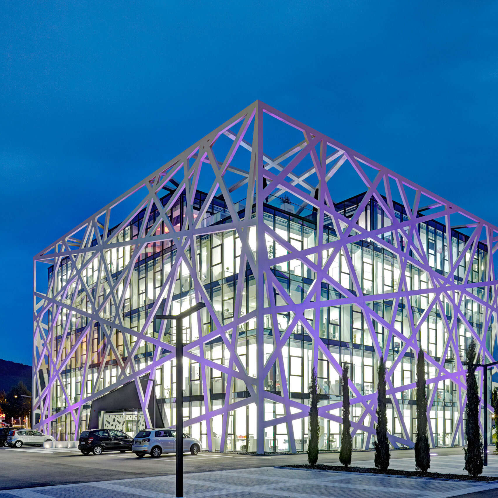 Exterior view of the QBIGone office building, a glass cube with a striking black and white steel mesh, illuminated at dusk