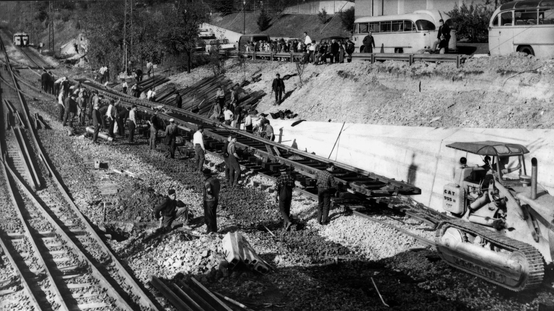 Arbeiter beim Gleisbau an der Geislinger Steige, unterstützt durch eine CAT-Baumaschine und beobachtet von Zuschauern im Hintergrund