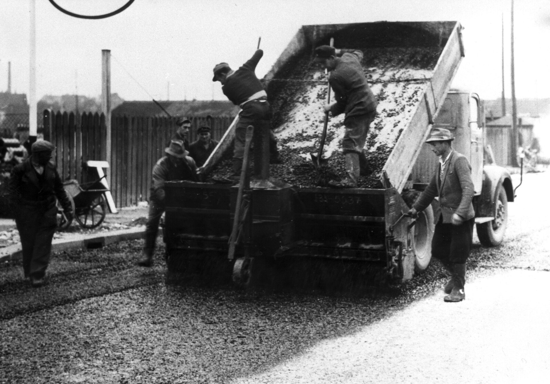 Schwarz-Weiß-Aufnahme von Straßenbauarbeiten um 1957: Arbeiter verteilen Kies von einem kippenden Lkw auf die Straße