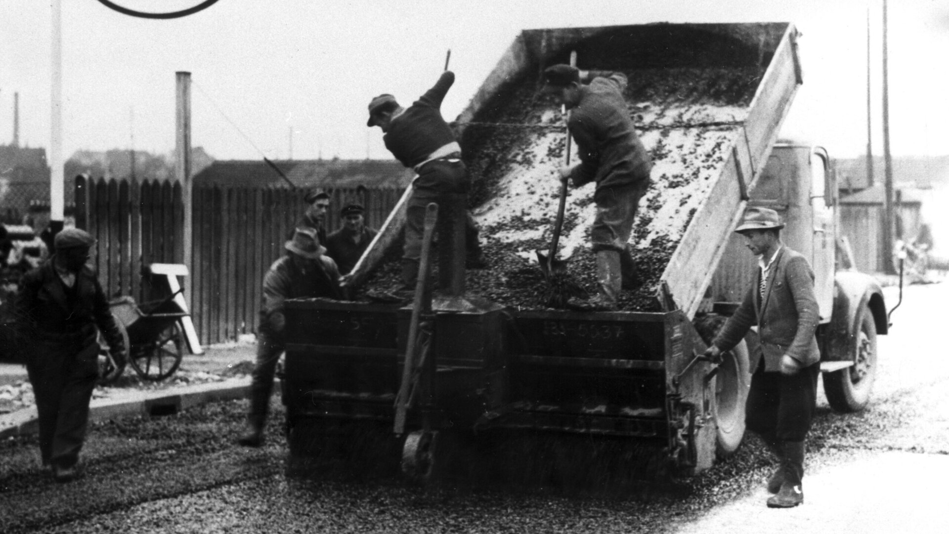 Schwarz-Weiß-Aufnahme von Straßenbauarbeiten um 1957: Arbeiter verteilen Kies von einem kippenden Lkw auf die Straße