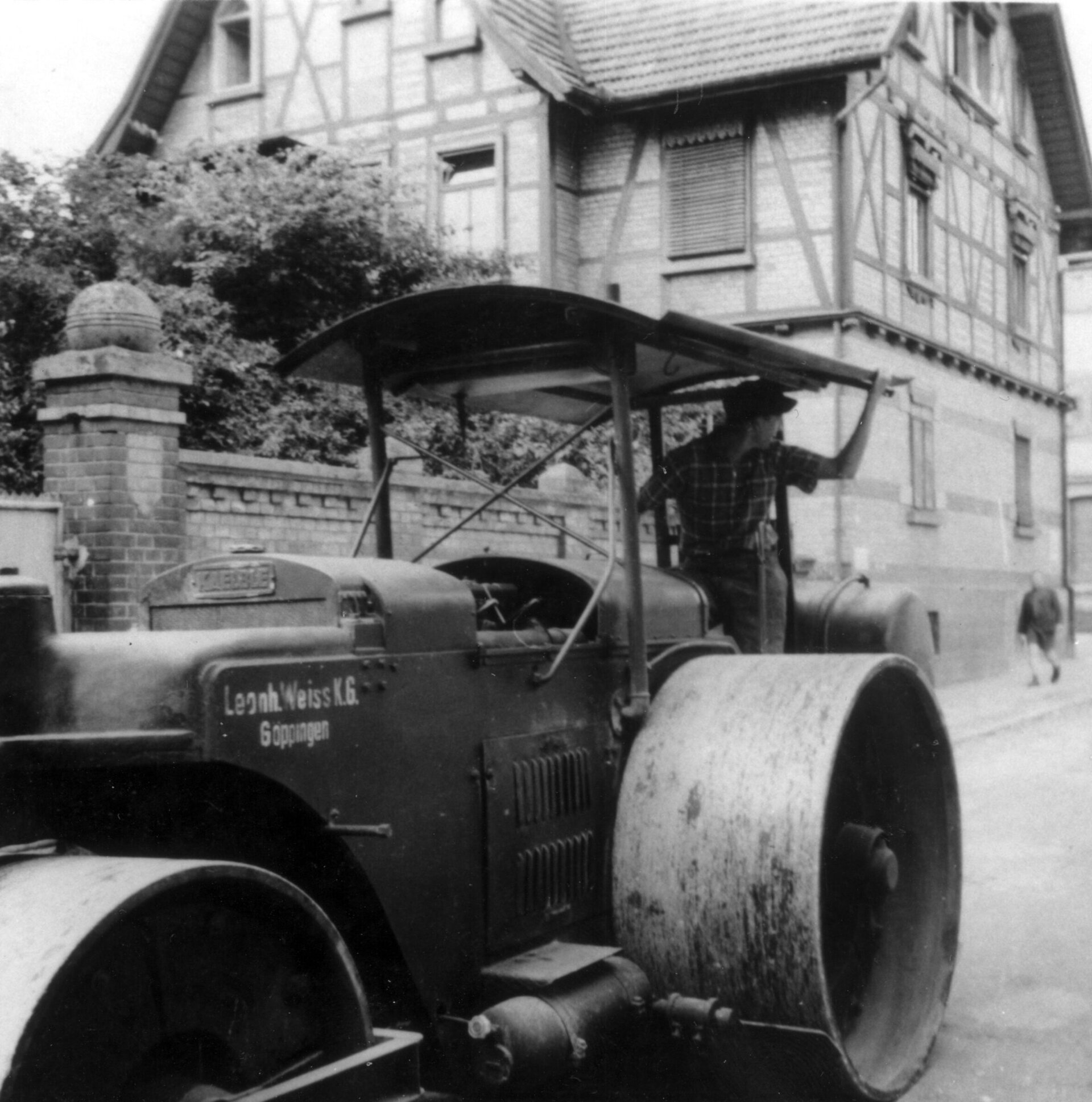 Schwarz-Weiß-Foto einer Walze mit der Aufschrift 'Leonhard Weiss KG Göppingen' im Einsatz beim Straßenbau um 1920, vor einem Fachwerkhaus