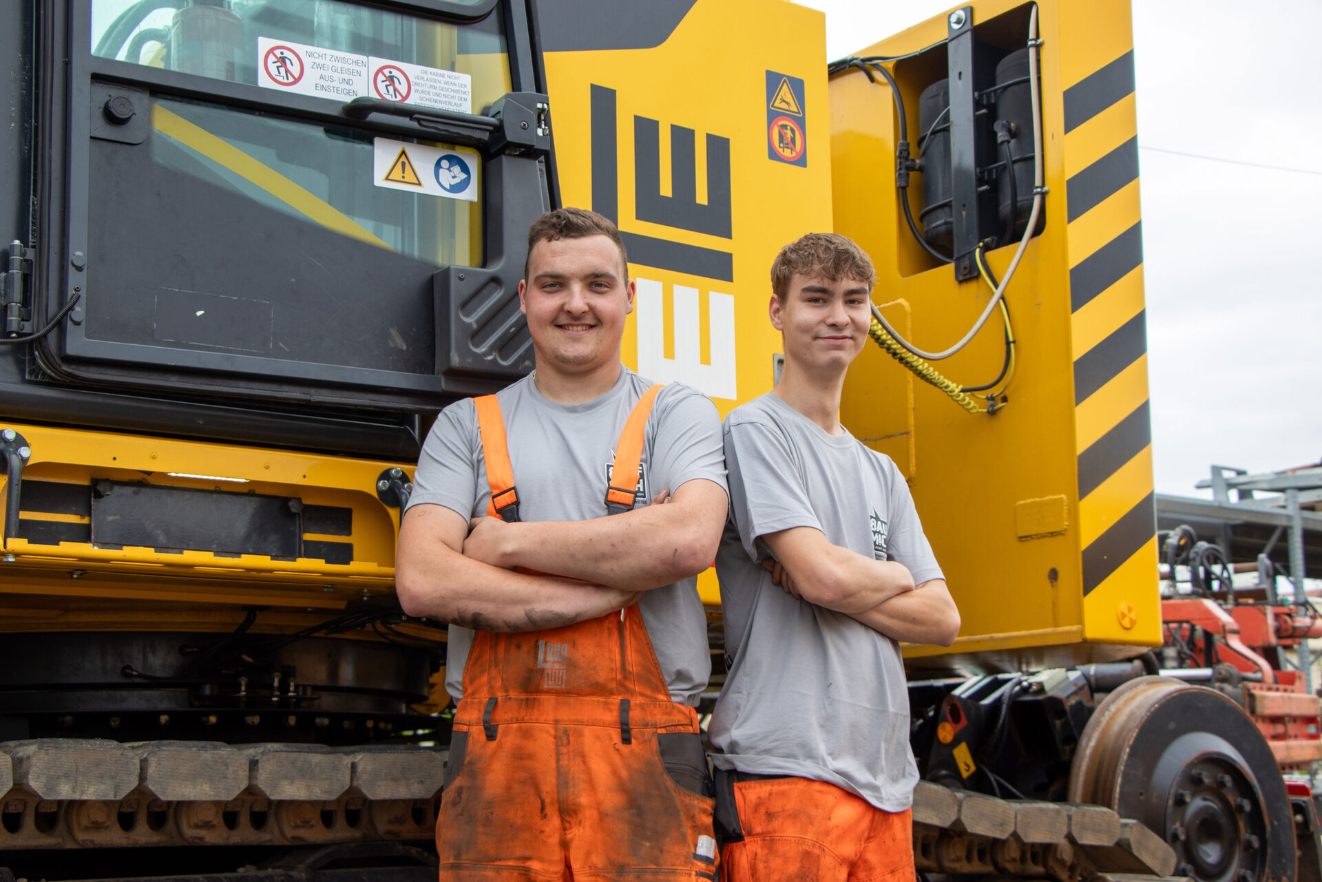 Zwei junge Männer in orangefarbener Arbeitskleidung posieren mit verschränkten Armen vor einer gelben Baumaschine mit LEONHARD WEISS Logo