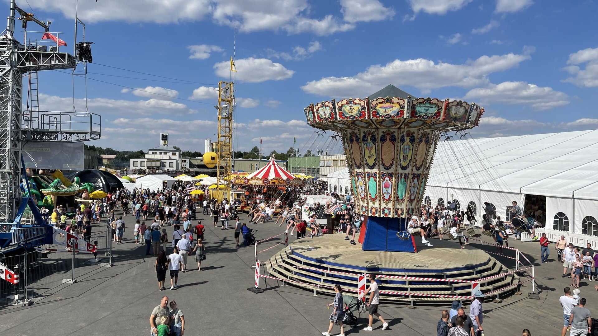 Belebtes Volksfest mit einem großen Kettenkarussell, Fahrgeschäften, Marktständen und zahlreichen Besuchern bei sonnigem Wetter.