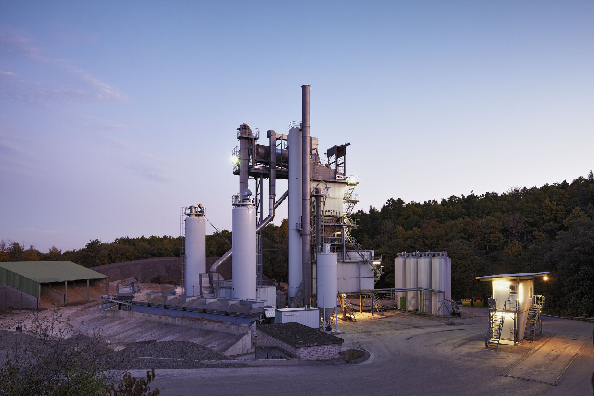 Asphaltmischanlage bei Dämmerung, umgeben von Wald, mit beleuchteten Silos und Produktionsgebäuden