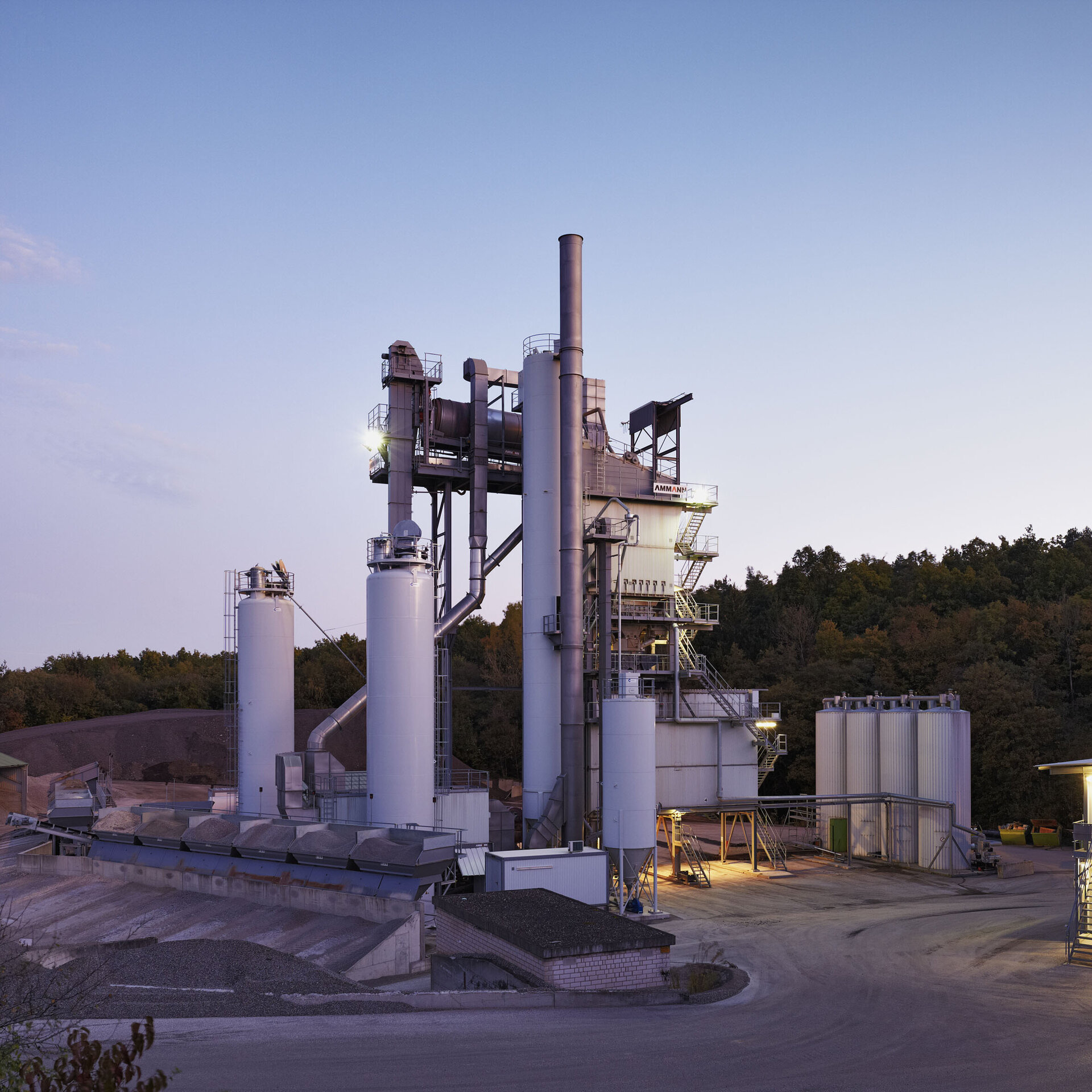 Asphalt mixing plant at dusk, surrounded by forest, with illuminated silos and production buildings