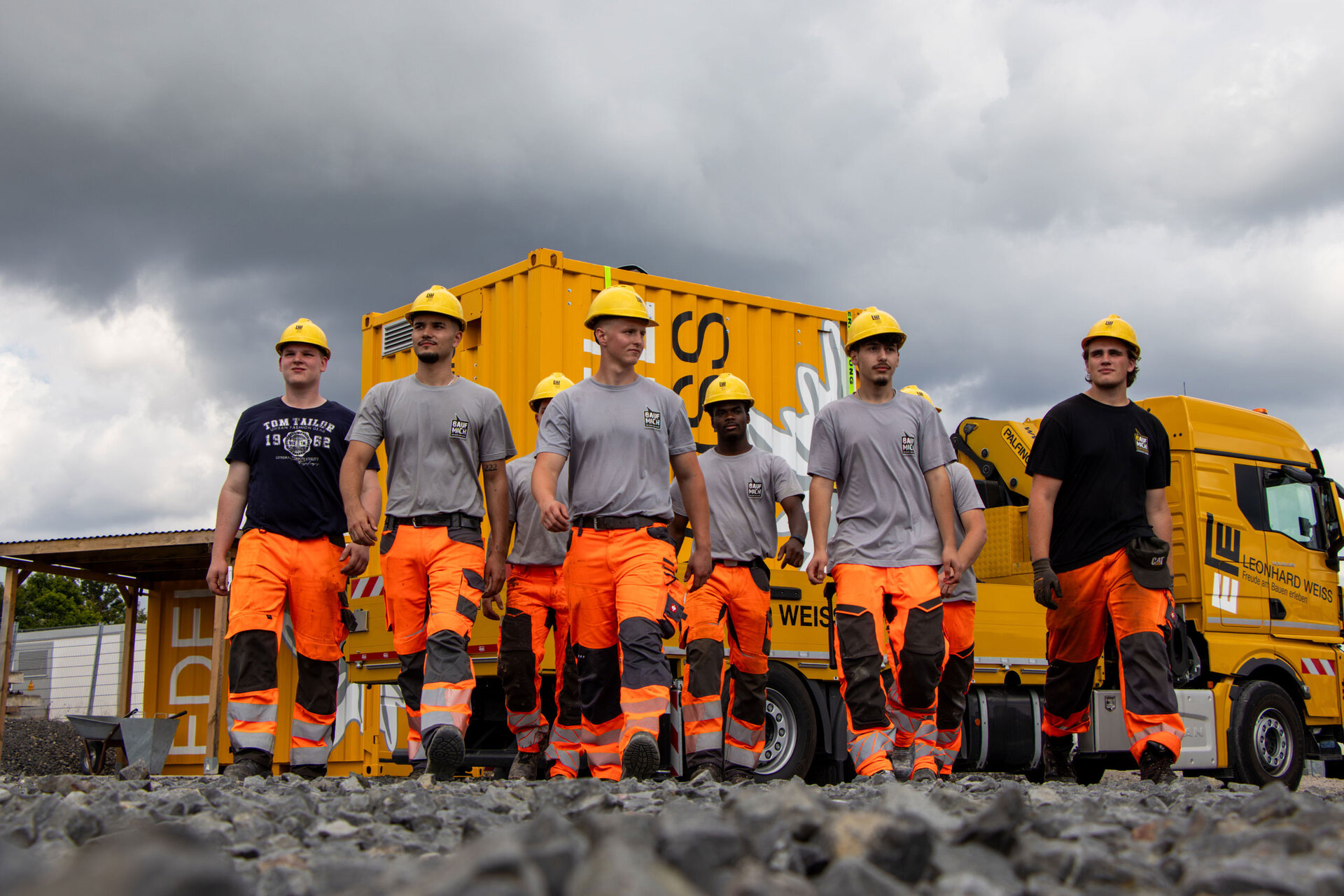 Gruppe junger Männer in orangefarbener Arbeitskleidung und gelben Schutzhelmen auf einer Baustelle, im Hintergrund ein gelber Container und ein LKW