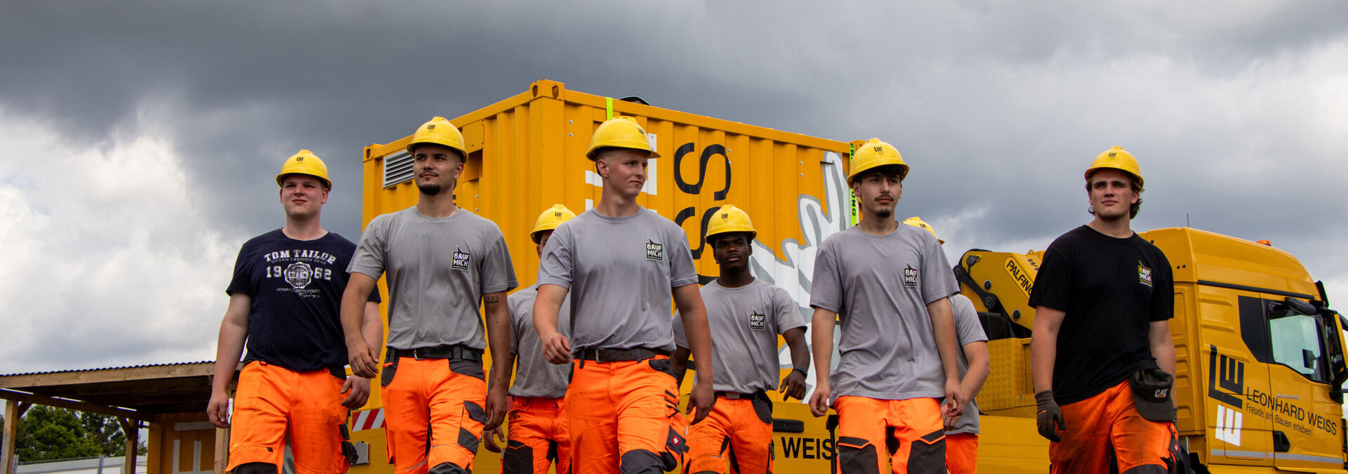 Gruppe junger Männer in orangefarbener Arbeitskleidung und gelben Schutzhelmen auf einer Baustelle, im Hintergrund ein gelber Container und ein LKW